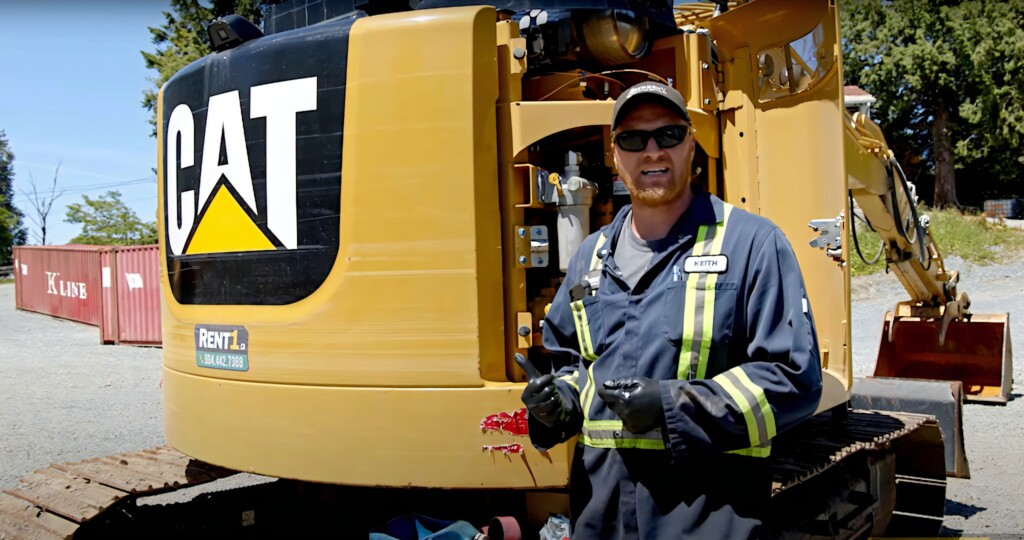 Muž v pracovnom odeve stojací pred stavebnou mašinou značky CAT.